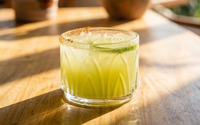 A green margarita in a cut-glass tumbler with a salted rim and lime wheel, in afternoon light by the window