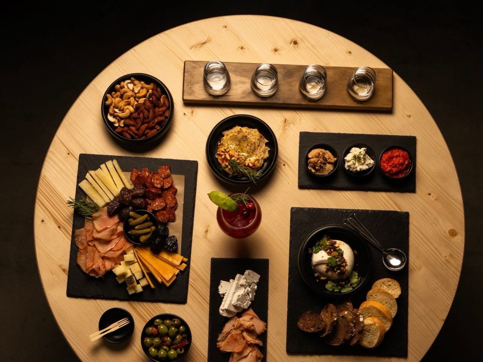 Boards of cured meats, cheeses, olives, hummus and burrata on a round wooden table, with a rakija flight