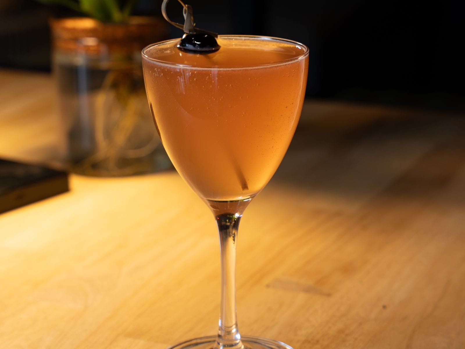 A pink cocktail in a nick-and-nora glass with a cherry, on the wooden bar
