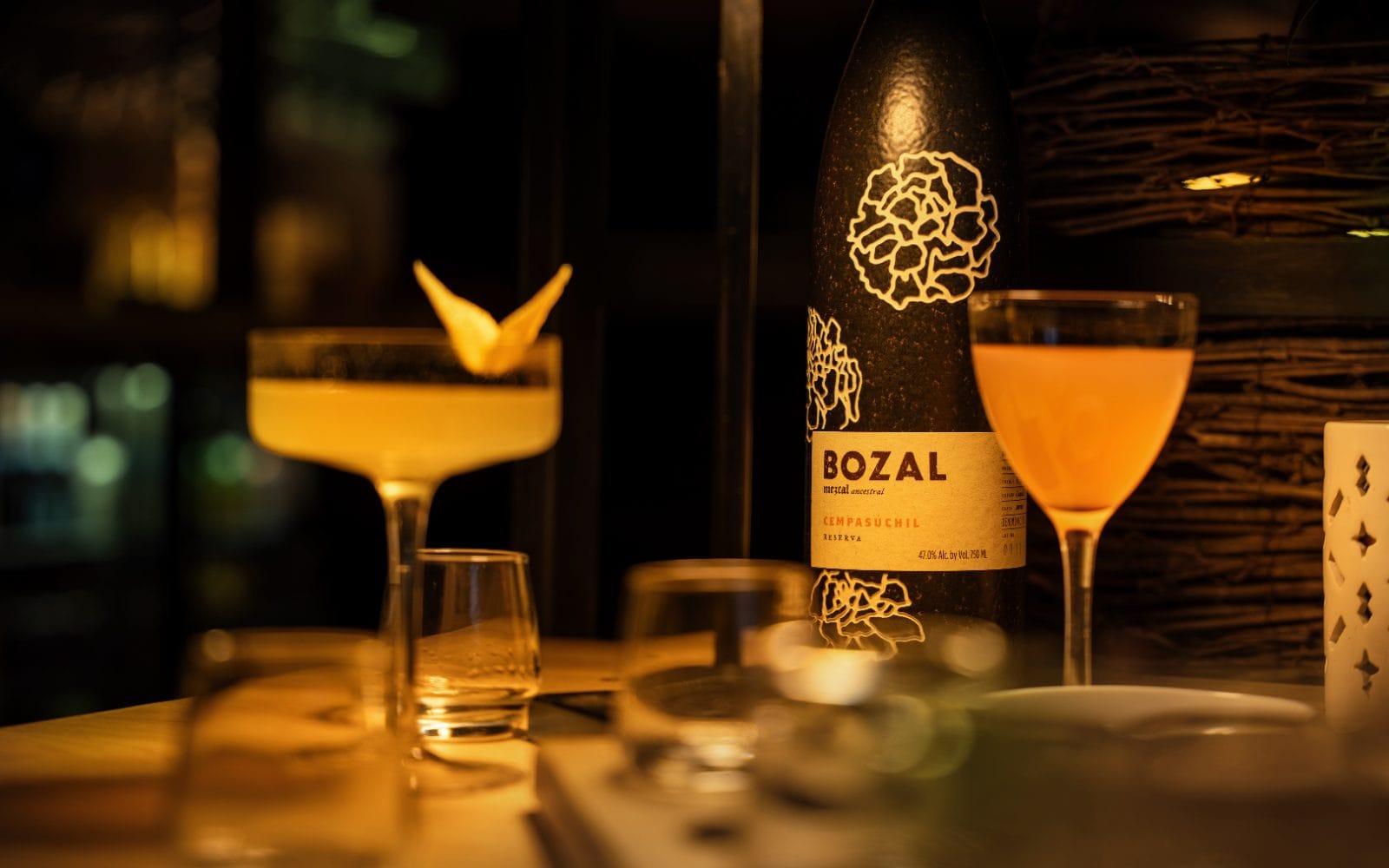 A bottle of Bozal Cempasúchil mezcal between a coupe and a nick-and-nora cocktail, in low light
